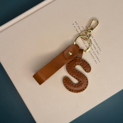 A tan leather keychain featuring a stitched letter 'S' charm and a matching leather strap, attached to gold-tone metal hardware.